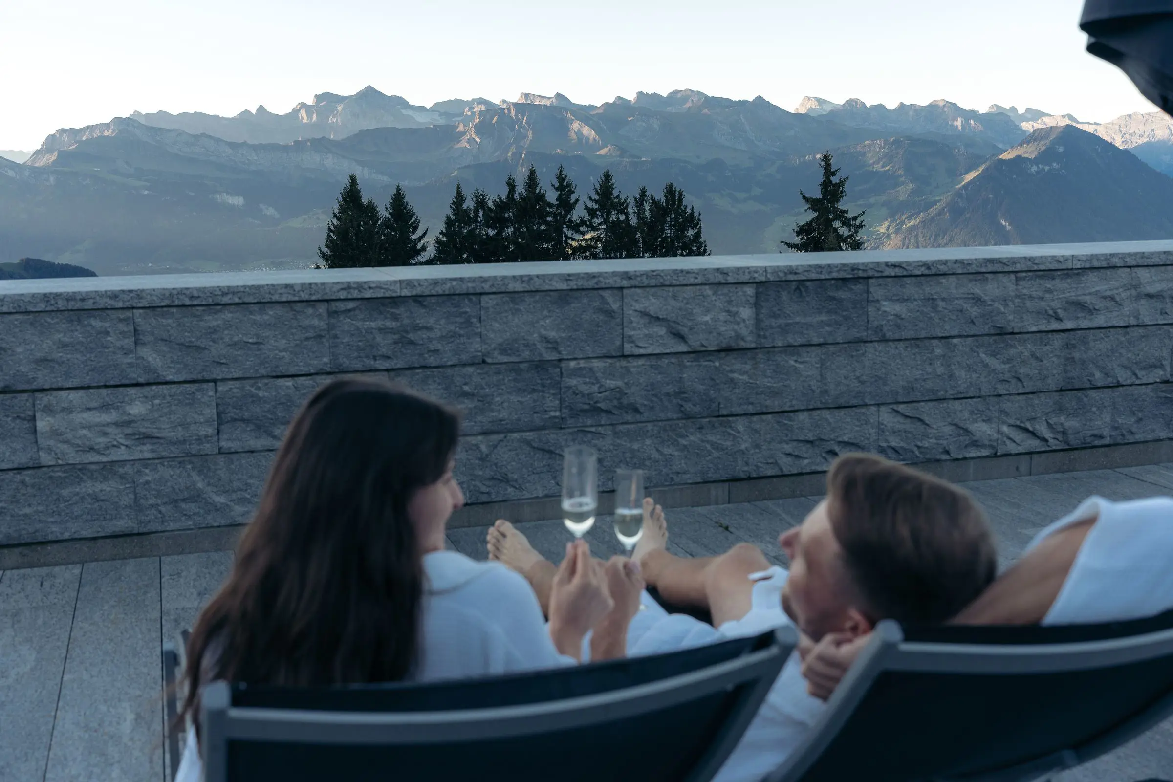 Zwei Gäste im Bademantel stossen auf der Terrasse des Mineralbads Rigi Kaltbad mit Blick auf die Alpen an